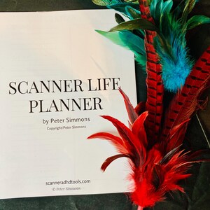 May include: A white spiral-bound Scanner Life Planner by Peter Simmons with black text on the cover, accompanied by a decorative arrangement of vibrant red, teal, and dark blue feathers with black striped patterns on the right side of the book.