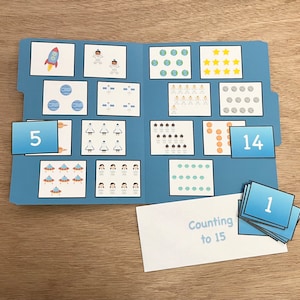 May include: A blue folder with a white card that says "Counting to 15". The folder has various images of astronauts, rockets, stars, and planets. There are also number cards with the numbers 1, 5, and 14.