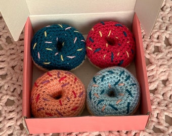 Puede incluir: Un conjunto de cuatro juguetes de ganchillo con forma de rosquilla dispuestos en una caja de cartón rosa. Las rosquillas son de color verde azulado, rosa brillante, melocotón y azul claro, con virutas de hilo bordadas en colores contrastantes.