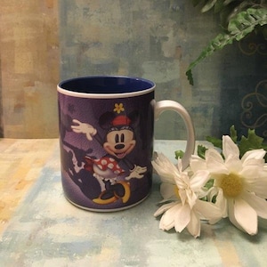 Puede incluir: Una taza de cer&aacute;mica azul y blanca con una imagen de Minnie Mouse con un vestido de lunares rojos y blancos y una flor amarilla en la cabeza. La taza tiene un borde y un asa azules.