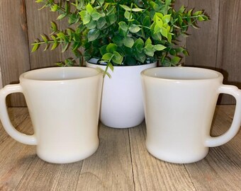 May include: Two matching off-white milk glass mugs with smooth, rounded bodies and C-shaped handles. The mugs feature a glossy, opaque finish and a simple, functional design. Each mug has a wide rim and a slightly tapered base, presenting a clean look.