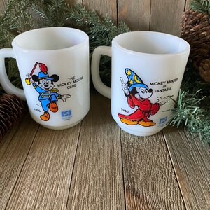 May include: Two white ceramic mugs with Mickey Mouse illustrations. The mug on the left features Mickey Mouse in a blue and red uniform with a drum and the text "The Mickey Mouse Club". The mug on the right features Mickey Mouse in a red and blue robe with a wizard hat and the text "Mickey Mouse in Fantasia".
