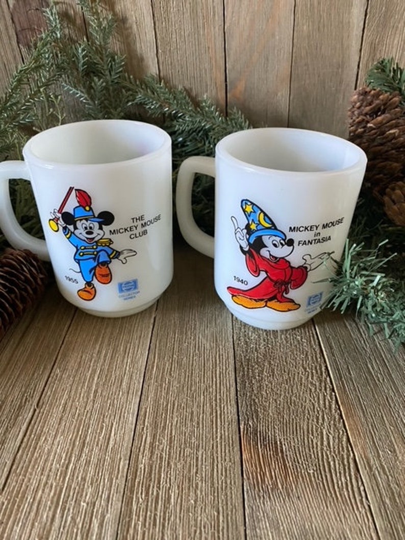 May include: Two white ceramic mugs with Mickey Mouse illustrations. The mug on the left features Mickey Mouse in a blue and red uniform with a drum and the text "The Mickey Mouse Club". The mug on the right features Mickey Mouse in a red and blue robe with a wizard hat and the text "Mickey Mouse in Fantasia".