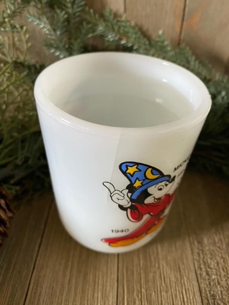 May include: White ceramic mug with a cartoon image of Mickey Mouse as a sorcerer. Mickey is wearing a blue hat with stars and a red robe. The mug has the text "Mickey Mouse" and "1940" printed on it.