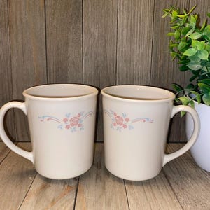 May include: Two off-white ceramic mugs with a floral design. The design features pink roses and blue accents. Each mug has a large handle and a simple, classic shape. The mugs are displayed on a wooden surface.