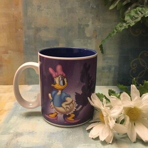 Puede incluir: Taza de cer&aacute;mica blanca con borde azul y fondo morado. La taza presenta una imagen de dibujos animados de Daisy Duck con un vestido azul y un lazo rosa. Su sombra est&aacute; detr&aacute;s de ella.