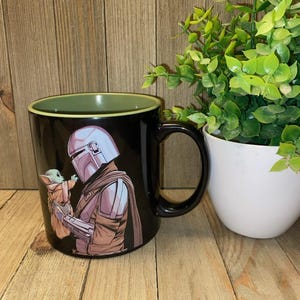 May include: Black ceramic mug featuring a detailed illustration of a character in a helmet holding a small green creature. The mug has a black handle and a green interior rim. A white pot with green plants is in the background.
