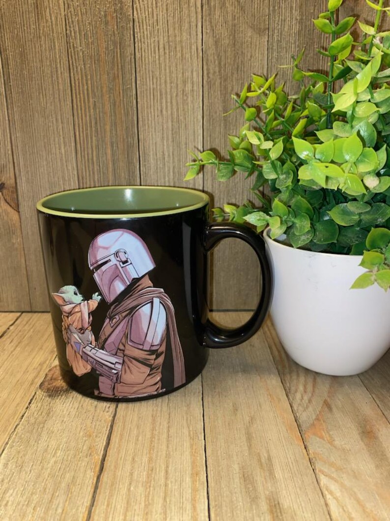 May include: Black ceramic mug featuring a detailed illustration of a character in a helmet holding a small green creature. The mug has a black handle and a green interior rim. A white pot with green plants is in the background.