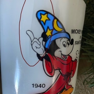 May include: White ceramic mug with a cartoon image of Mickey Mouse dressed as a wizard. Mickey is wearing a blue hat with stars and a crescent moon. The mug has the text "Mickey Mouse in Fantasia" and "1940" printed on it.