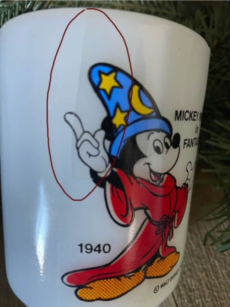 May include: White ceramic mug with a cartoon image of Mickey Mouse dressed as a wizard. Mickey is wearing a blue hat with stars and a crescent moon. The mug has the text "Mickey Mouse in Fantasia" and "1940" printed on it.
