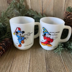 May include: Two white ceramic mugs with images of Mickey Mouse. The mug on the left features Mickey Mouse in a blue and red uniform with the text "The Mickey Mouse Club" and "1955". The mug on the right features Mickey Mouse in a red robe with a wizard hat and the text "Mickey Mouse in Fantasia" and "1940".