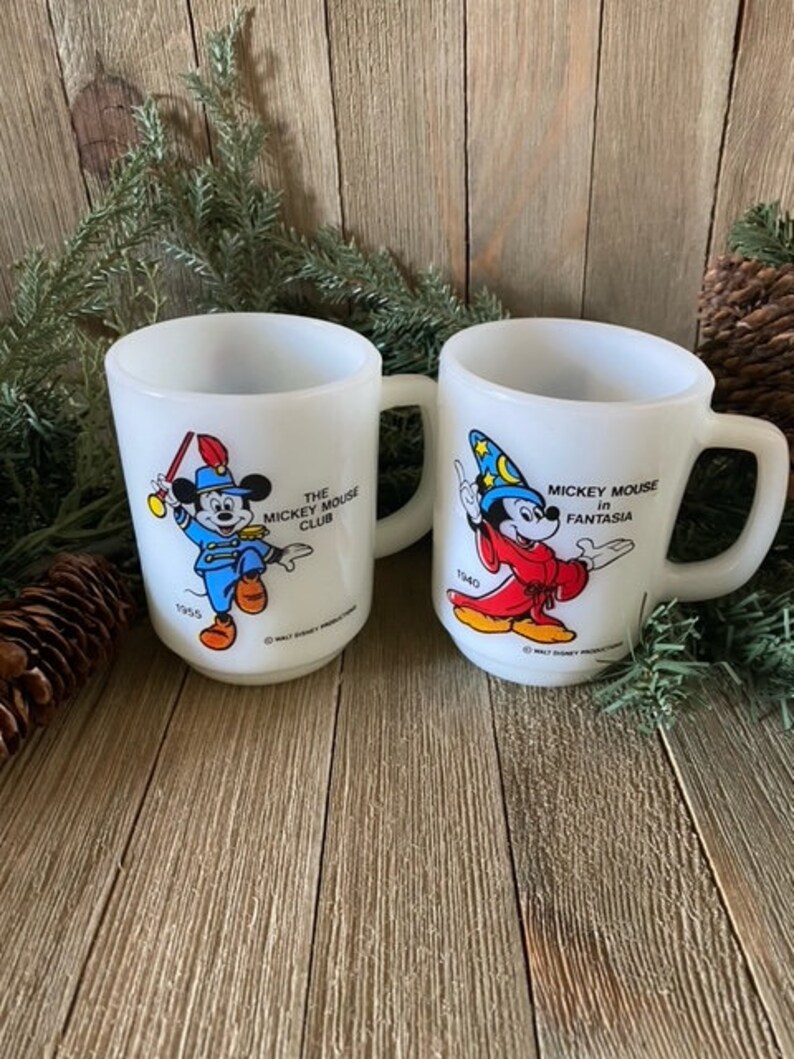 May include: Two white ceramic mugs with images of Mickey Mouse. The mug on the left features Mickey Mouse in a blue and red uniform with the text "The Mickey Mouse Club" and "1955". The mug on the right features Mickey Mouse in a red robe with a wizard hat and the text "Mickey Mouse in Fantasia" and "1940".