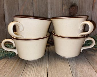 May include: A set of four cream-colored ceramic cups with speckled textures and dark brown rims. Each cup features a curved handle and a wide, flared opening. The cups are arranged in two stacks, with two cups positioned on the left and two on the right.
