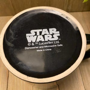 May include: Black ceramic mug with the Star Wars logo in white. The bottom of the mug features the text "Dishwasher and Microwave Safe" and "Made in China". The mug has a black interior and a white rim.