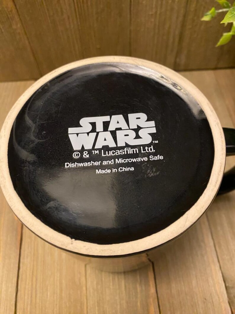 May include: Black ceramic mug with the Star Wars logo in white. The bottom of the mug features the text "Dishwasher and Microwave Safe" and "Made in China". The mug has a black interior and a white rim.