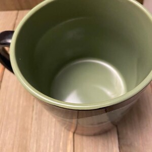 May include: A green ceramic mug with a black handle. The interior and rim are a lighter shade of green. The mug sits on a wooden surface.