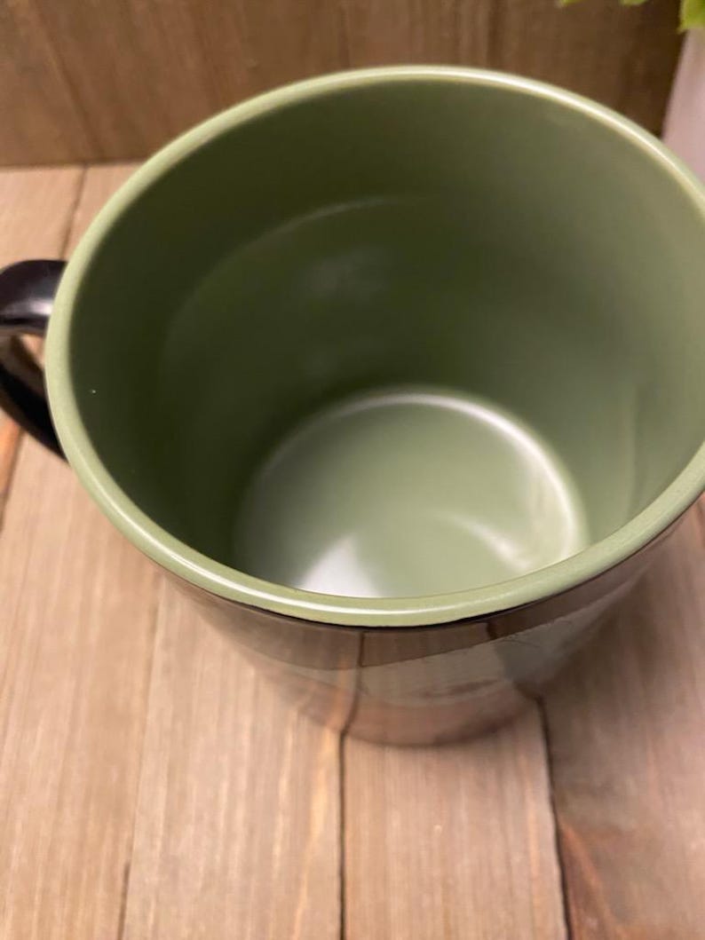 May include: A green ceramic mug with a black handle. The interior and rim are a lighter shade of green. The mug sits on a wooden surface.