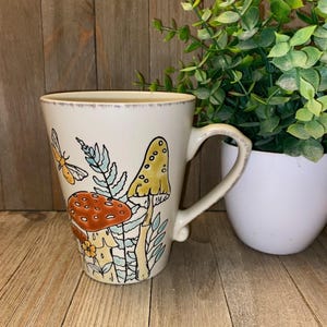 May include: A cream-colored ceramic mug featuring a decorative illustration of orange and yellow mushrooms, green ferns, and a small moth. The mug has a tapered shape, a curved handle, and a distressed, weathered finish along the rim and handle.