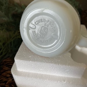 May include: White ceramic pitcher with a round handle and a white, opaque glaze. The bottom of the pitcher has a circular label that reads "OVEN-PROOF 34 MADE IN USA".