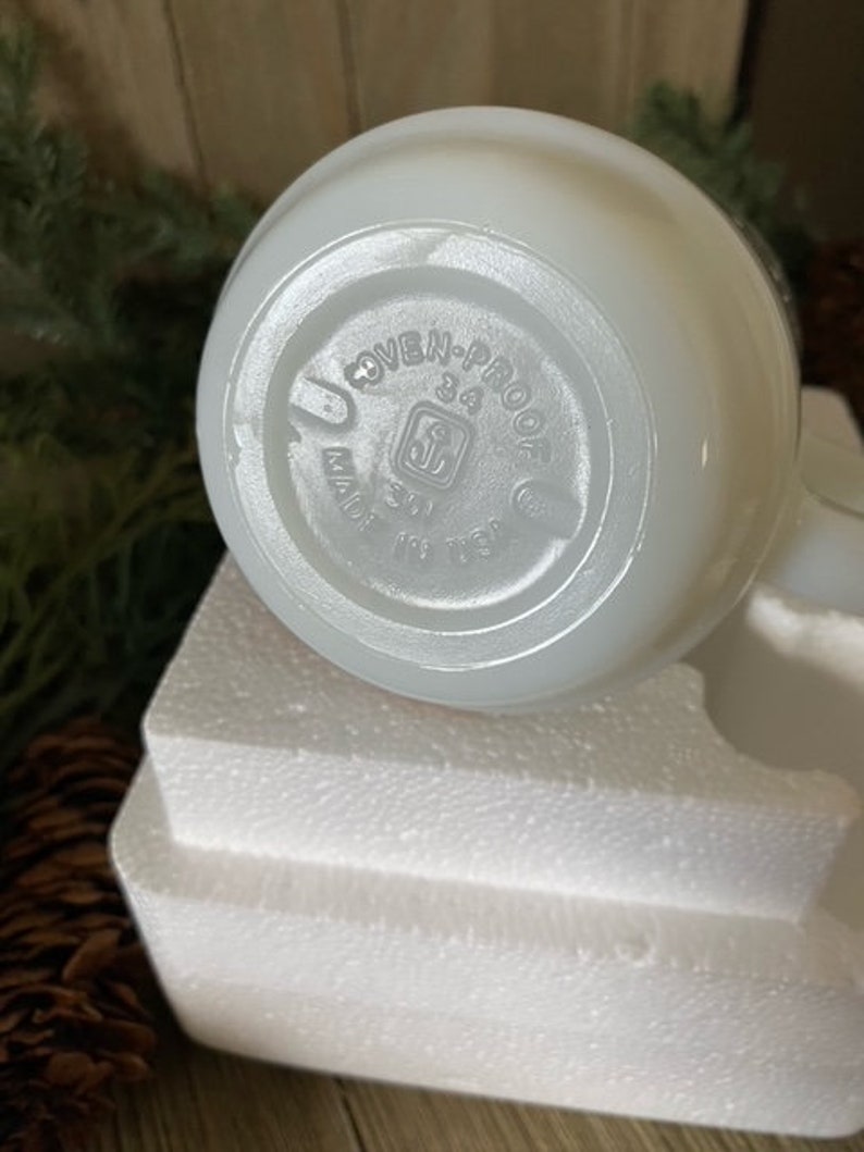 May include: White ceramic pitcher with a round handle and a white, opaque glaze. The bottom of the pitcher has a circular label that reads "OVEN-PROOF 34 MADE IN USA".