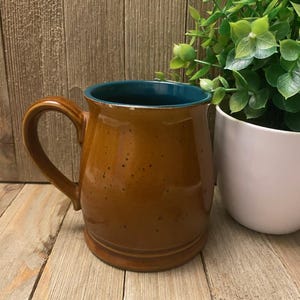 May include: A brown ceramic mug with a teal interior. The mug has a handle and a speckled finish. A white potted plant with green leaves is next to the mug. The mug is on a wooden surface.
