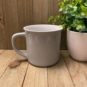 May include: A light gray ceramic mug with a curved handle. The mug sits on a light brown wooden surface. A white planter with green foliage is to the right of the mug.