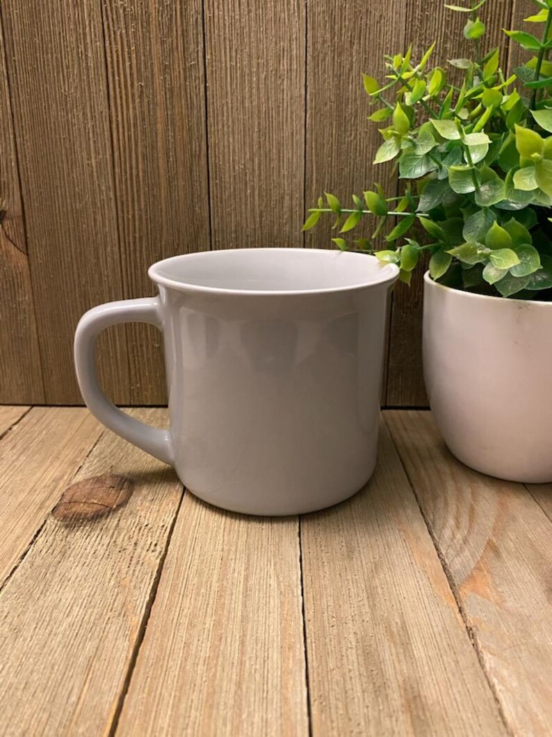 May include: A light gray ceramic mug with a curved handle. The mug sits on a light brown wooden surface. A white planter with green foliage is to the right of the mug.