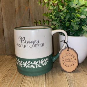 May include: A two-toned ceramic mug with a white upper section featuring the grey text Prayer changes things and a dark green lower section with a white leaf pattern. A brown scalloped paper tag reads Seeds & Sunshine in black text.
