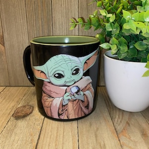 May include: Black ceramic mug featuring a green-skinned character with large ears, holding a blue sphere. The character is wearing a tan robe. The mug has a green interior rim and a black handle. The background is a wooden surface.