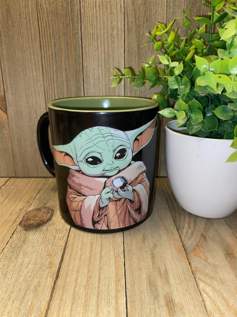 May include: Black ceramic mug featuring a green-skinned character with large ears, holding a blue sphere. The character is wearing a tan robe. The mug has a green interior rim and a black handle. The background is a wooden surface.