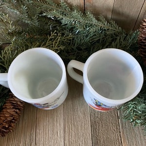 May include: Two white ceramic mugs with colorful illustrations. The mugs have handles and are sitting on a wooden surface.