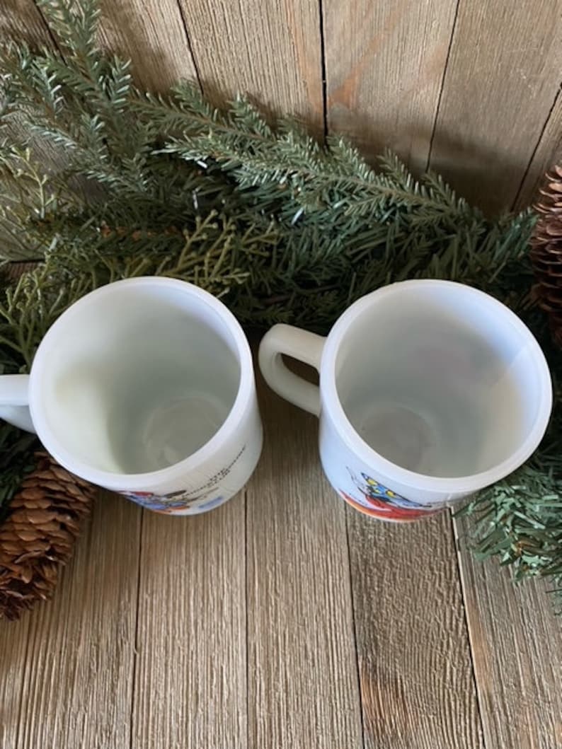 May include: Two white ceramic mugs with colorful illustrations. The mugs have handles and are sitting on a wooden surface.