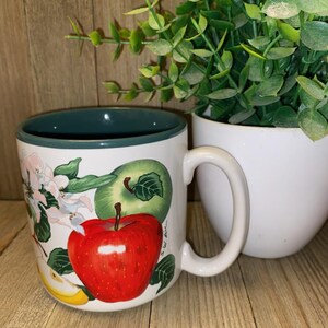 May include: A white ceramic mug featuring a painted design of a bright red apple, a green apple, and light pink apple blossoms. The mug has a dark green interior and a white handle. The exterior surface displays a glossy finish with detailed fruit illustrations.