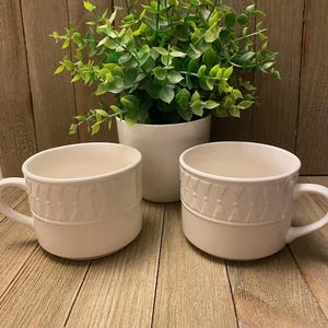 May include: Two white ceramic mugs with a decorative pattern around the top rim and a handle. A small white potted plant with green leaves sits behind the mugs on a wooden surface. The mugs are designed for hot beverages.