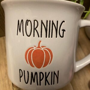May include: White ceramic mug with the words "MORNING PUMPKIN" in black lettering and an orange pumpkin graphic. The mug has a curved handle and a slightly flared rim. A perfect addition to any fall-themed kitchen.