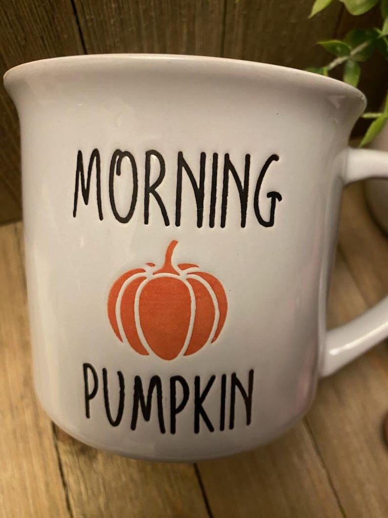 May include: White ceramic mug with the words "MORNING PUMPKIN" in black lettering and an orange pumpkin graphic. The mug has a curved handle and a slightly flared rim. A perfect addition to any fall-themed kitchen.