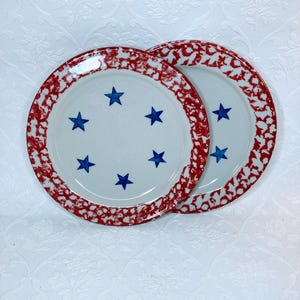 May include: Two white ceramic plates with a red speckled border and blue star designs. The plates are round and have a classic, vintage aesthetic. The plates are likely for serving food.