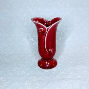 May include: A red ceramic vase with a flared top and a pedestal base. The vase has a glossy finish and a decorative swirl design on the sides. The vase is approximately 7 inches tall.