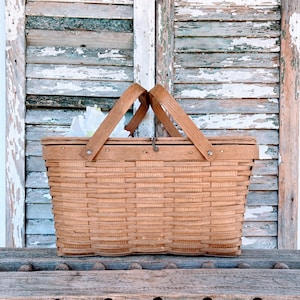 May include: A vintage wicker picnic basket with a brown wooden handle and a latch closure. The basket is sitting on a weathered wooden surface.
