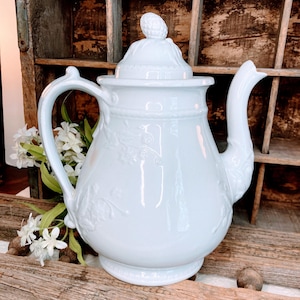 May include: A white porcelain teapot with a floral design. The teapot has a lid with a knob in the shape of a flower bud. The teapot is sitting on a wooden surface.