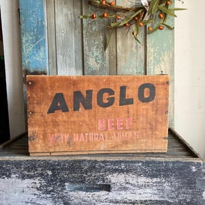 May include: A weathered wooden box with the word "ANGLO" in large black letters and "BEEF" in pink letters. The text "WITH NATURAL JUICES" is also visible. The box is sitting on a rustic, weathered wooden surface.