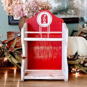 May include: A distressed, red wooden shelf with two tiers and a decorative top. The top features a white graphic of a flower in a vase. The shelf is painted with a distressed, vintage look, and is perfect for displaying small items.