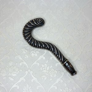 May include: A silver-toned, curved handle with a spiral design at the top and a textured, cylindrical base. The handle has a twisted, ridged pattern along its length. The object is likely a decorative element or part of a larger item.