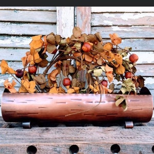 May include: A copper-colored planter shaped like a log with a rustic wood grain texture. The planter is filled with dried fall foliage in shades of orange, brown, and yellow.