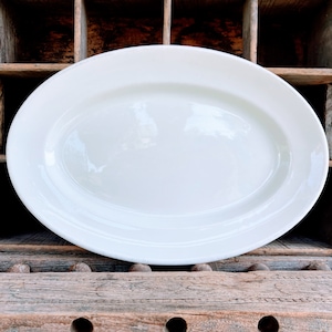 May include: A large, white oval serving platter with a slightly raised rim. The platter is made of ceramic and has a glossy finish. The background is a rustic wooden surface.