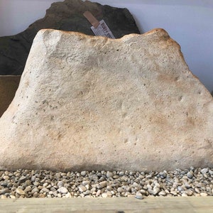 May include: A large, light brown, irregularly shaped rock with a smooth surface. The rock is sitting on a bed of small, gray pebbles.
