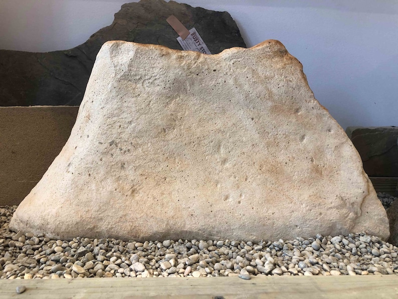 May include: A large, light brown, irregularly shaped rock with a smooth surface. The rock is sitting on a bed of small, gray pebbles.
