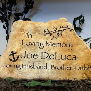 May include: A light brown stone with the inscription "In Loving Memory Joe DeLuca Loving Husband, Brother, Father" and a small anchor symbol.