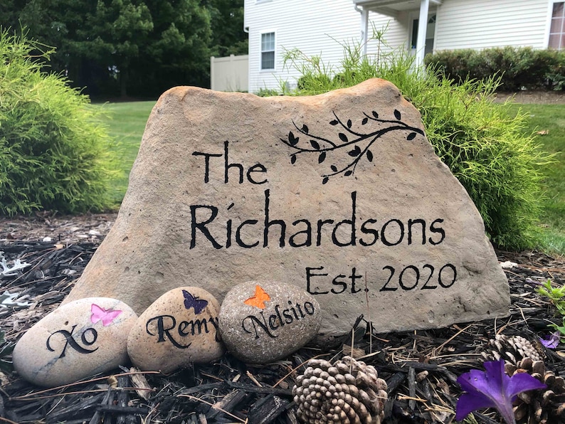 May include: A large, light brown rock with the words "The Richardsons Est. 2020" painted in black.  Three smaller rocks with painted butterflies and names are in front of the large rock.