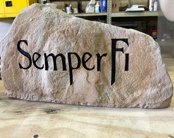 May include: A light-coloured stone with the words "Semper Fi" in black lettering. The stone has a rough, textured surface. It is likely used as a decorative item or sign, possibly for a garden.